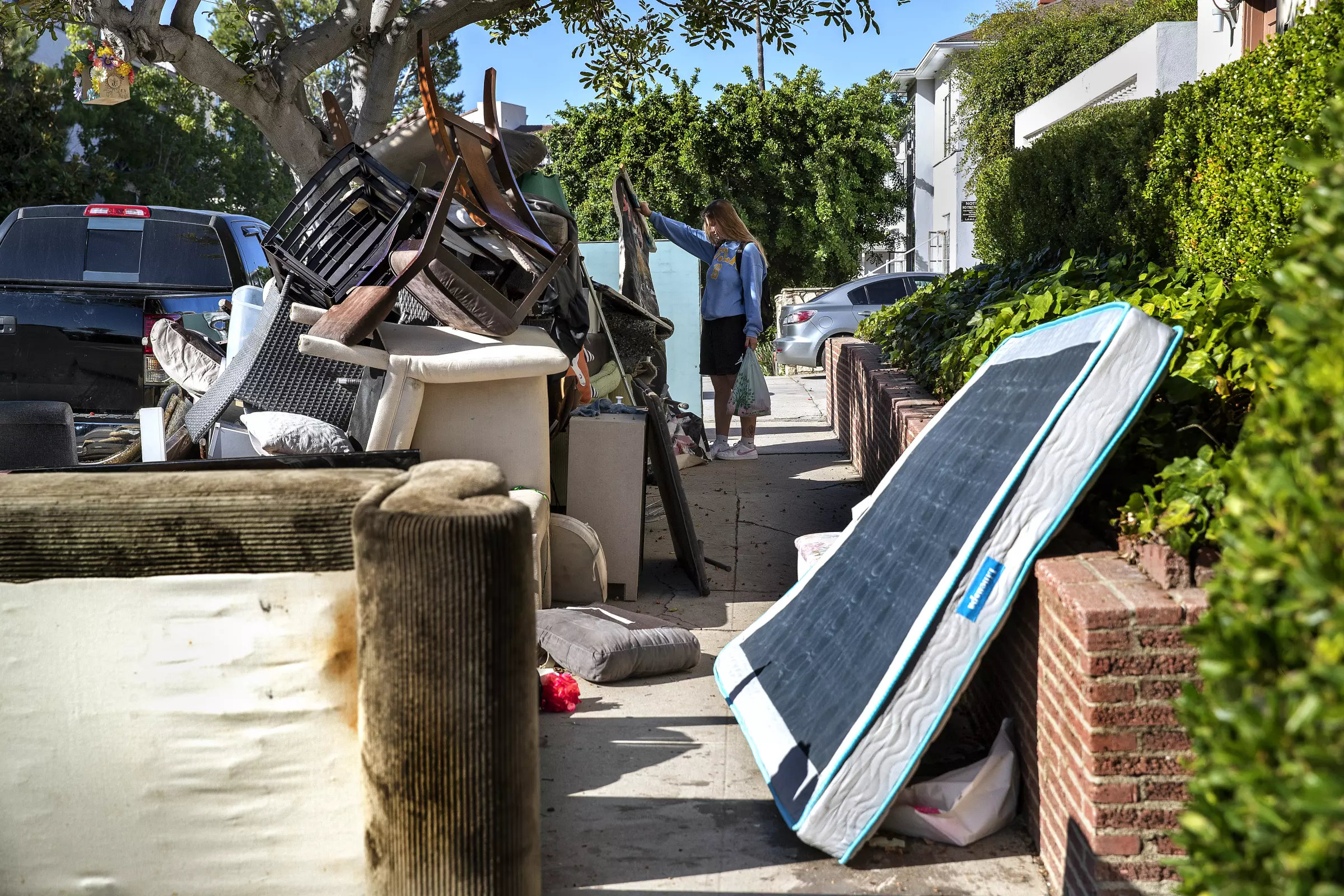 Piles of trash on the curb from college students on move out day. A woman is looking at a rug from what was left behind.